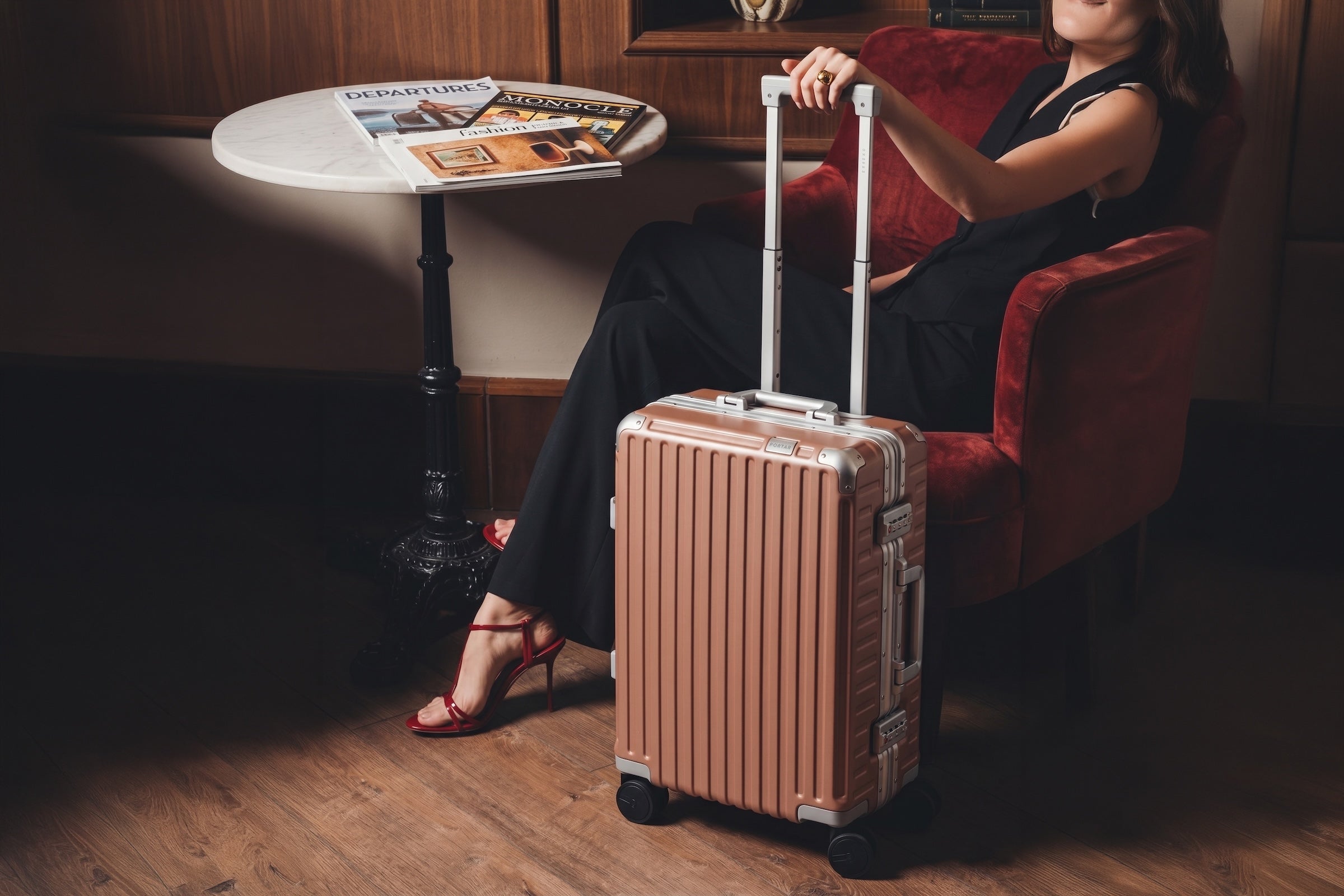 A seated traveler rests a hand on the extended handle of a rose-toned aluminum Portar carry-on beside a marble cafe table in a warm lounge.