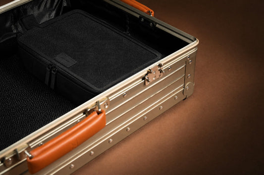 A black mesh-top packing cube inside an open aluminum Portar carry-on with a leather handle, on a warm brown backdrop.