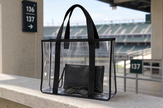 A clear stadium-approved tote with black trim, an interior pouch, and sunglasses rests on a concourse ledge near the seating sections.