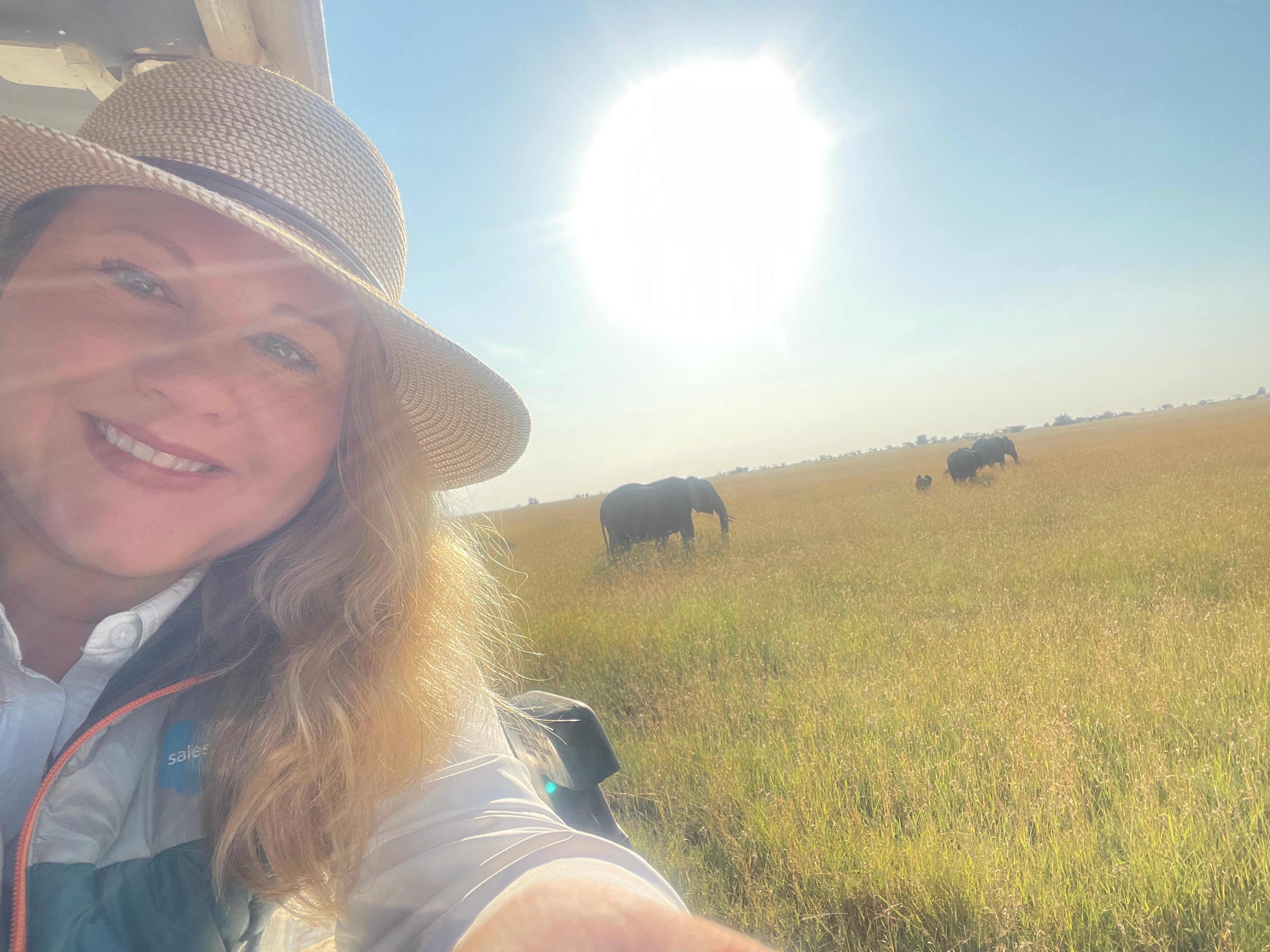 Smiling traveler in a sun hat on safari, with elephants grazing across the golden savanna of Masai Mara, Kenya.