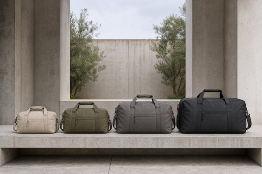 Four travel duffles in graduated sizes lined up smallest to largest on a concrete bench against architectural concrete and olive trees.