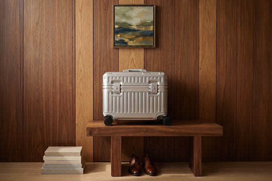 A silver aluminum Portar Concord pilot carry-on stands on a walnut bench against wood paneling, with leather dress shoes beneath and framed art above.