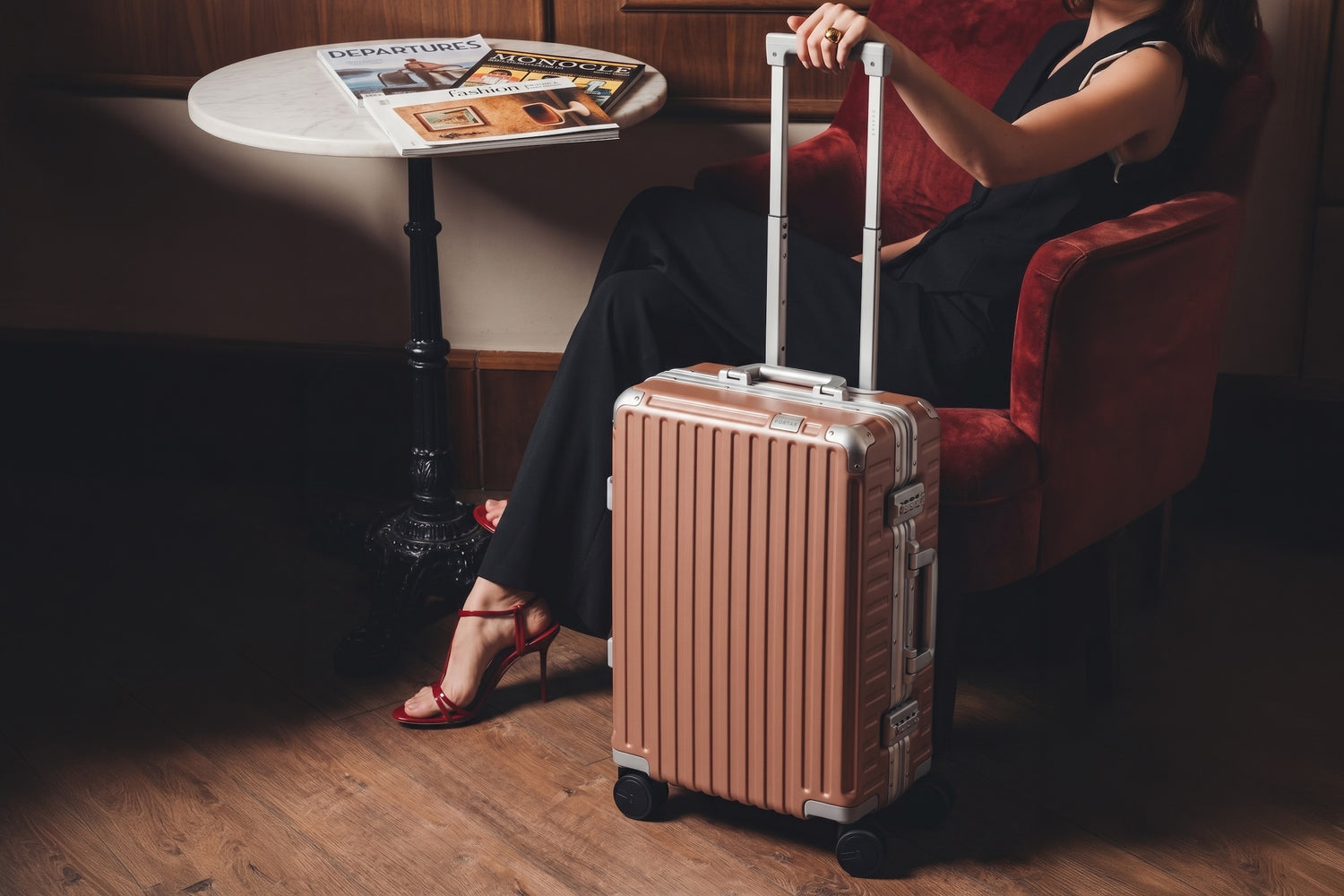 A seated traveler rests a hand on the extended handle of a rose-toned aluminum Portar carry-on beside a marble cafe table in a warm lounge.
